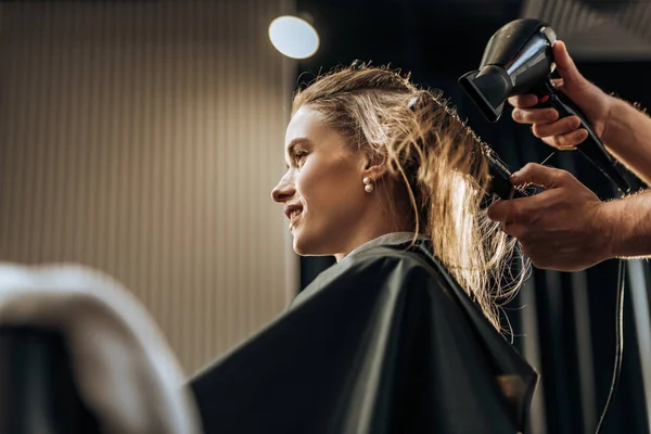 Recortado disparo de peluquero secando el pelo a la chica sonriente en salón de belleza - foto de stock