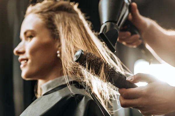 Gros plan vue partielle de coiffeur peignage et séchage des cheveux à belle jeune femme dans le salon de beauté — Photo de stock