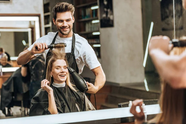 Sonriente joven peluquero peinando y secando el cabello a la joven feliz en el salón de belleza - foto de stock