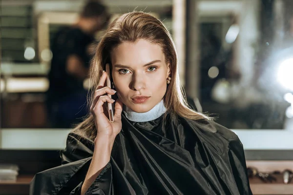 Atractiva mujer joven hablando por teléfono inteligente y mirando a la cámara en el salón de belleza - foto de stock