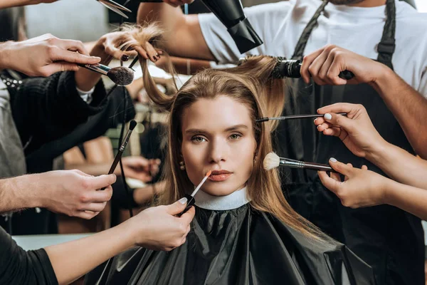 Belle fille regardant caméra tandis que les stylistes appliquant maquillage et faire coiffure dans le salon de beauté — Photo de stock
