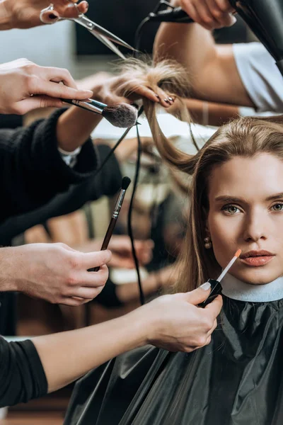 Chica atractiva mirando a la cámara mientras estilistas aplicando maquillaje y peinado - foto de stock