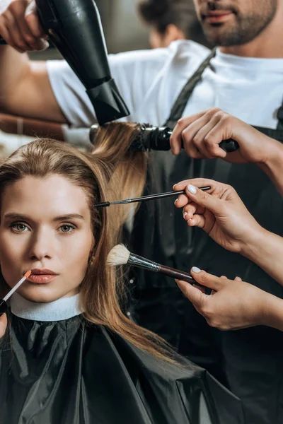 Menina bonita olhando para a câmera enquanto estilistas aplicando maquiagem e fazendo penteado — Fotografia de Stock