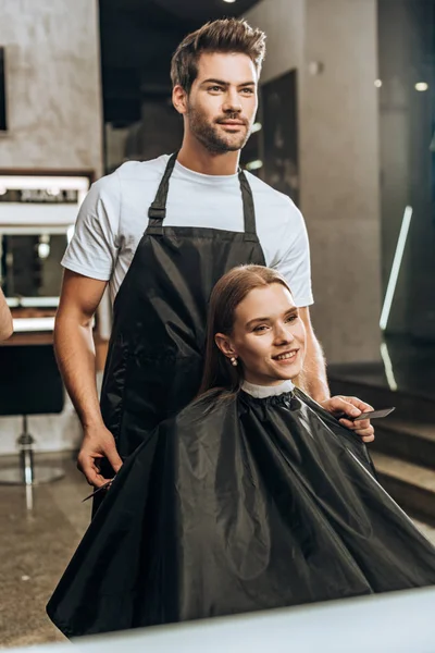 Junge Friseurin hält Kamm mit Schere und schaut Spiegel mit schöner Frau im Schönheitssalon — Stockfoto