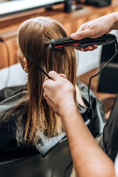 Cropped shot of hairstylist holding comb and using hair straightener in beauty salon — Stock Photo