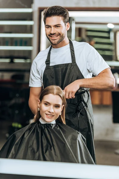 Улыбающийся парикмахер с помощью выпрямителя для волос и счастливая девушка, смотрящая на зеркало в салоне красоты — стоковое фото