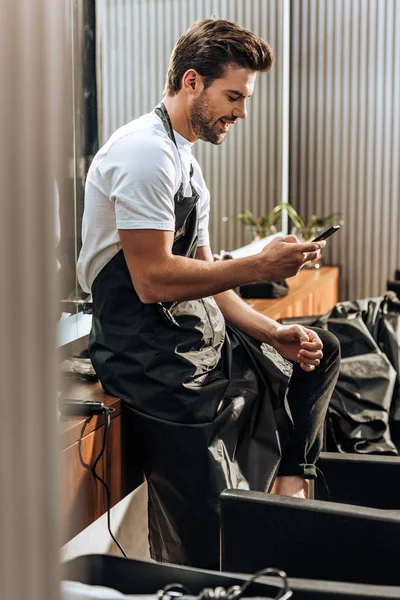 Side view of smiling young hairstylist in apron using smartphone in beauty salon — Stock Photo