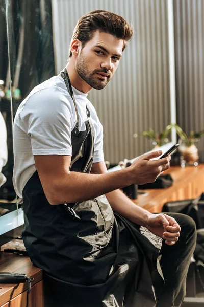 Guapo joven peluquero usando teléfono inteligente y mirando a la cámara en el salón de belleza - foto de stock