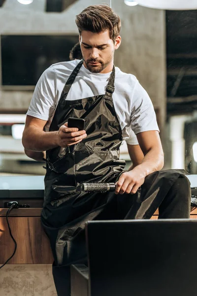 Beau jeune coiffeur tenant une brosse à cheveux et utilisant un smartphone dans un salon de beauté — Photo de stock