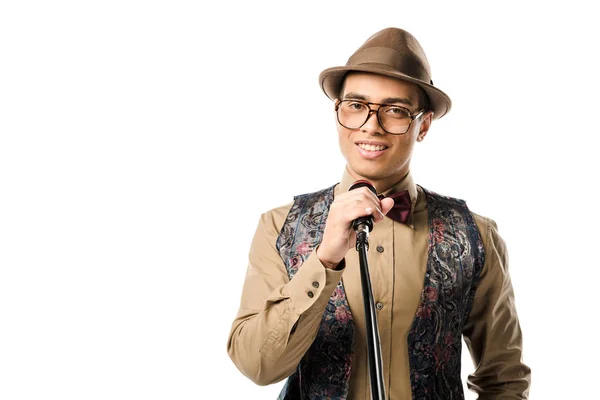 Sonriente hombre de raza mixta en elegante sombrero y anteojos cantando en micrófono aislado en blanco - foto de stock