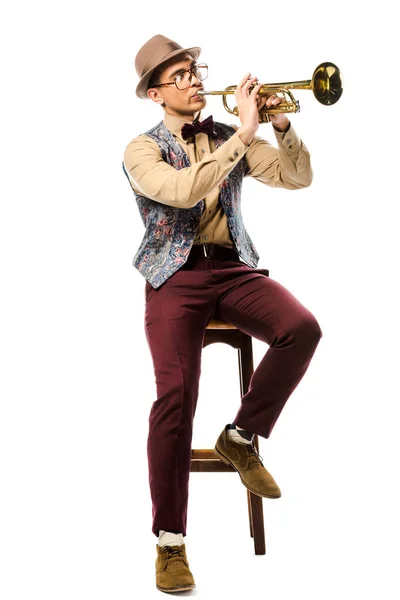Stylish hipster man in hat and eyeglasses playing on trumpet while sitting on chair isolated on white — Stock Photo
