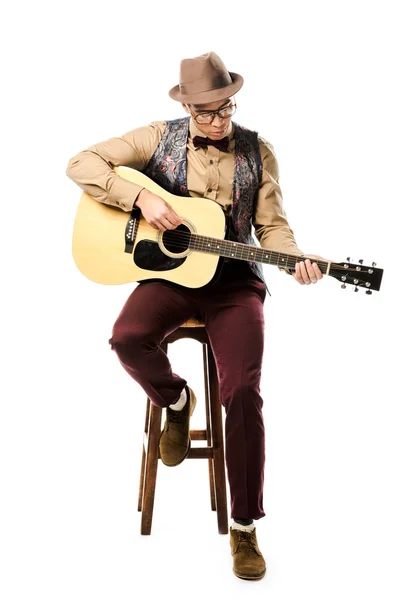 Fokussierte gemischte Rasse männliche Musiker mit Hut und Brille spielen auf akustischer Gitarre, während sie auf Stuhl isoliert auf weiß sitzen — Stockfoto