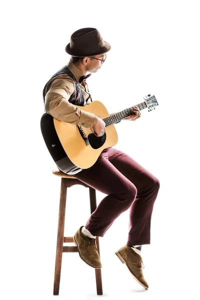 Joven músico masculino de raza mixta con sombrero y anteojos tocando la guitarra acústica mientras está sentado en una silla aislada en blanco - foto de stock