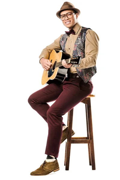 Alegre mestizo de raza masculina con sombrero y gafas tocando en la guitarra acústica mientras está sentado en silla aislado en blanco - foto de stock