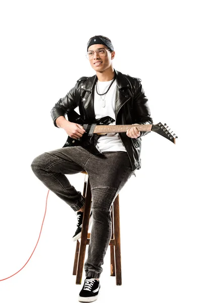 Sonriente músico de rock masculino en chaqueta de cuero tocando la guitarra eléctrica mientras está sentado en la silla aislado en blanco - foto de stock