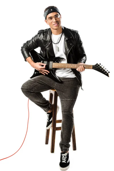 Cheerful male rock musician in leather jacket playing on electric guitar while sitting on chair isolated on white — Stock Photo