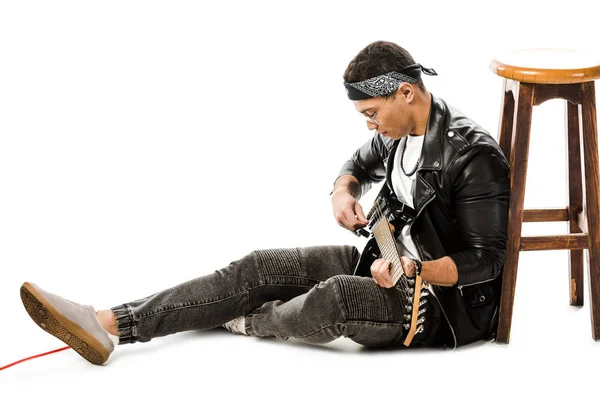 Концентрированный молодой человек музыкант в кожаной куртке, играющий на электрогитаре, сидя на полу возле стула, изолированного на белом — стоковое фото