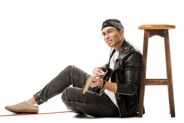 Male rock musician in leather jacket playing on electric guitar while sitting on floor near chair isolated on white — Stock Photo