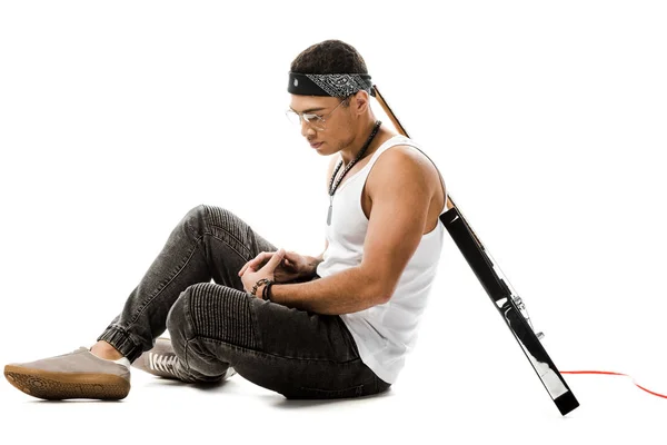 Young male rock musician sitting on floor with black electric guitar on back isolated on white — Stock Photo