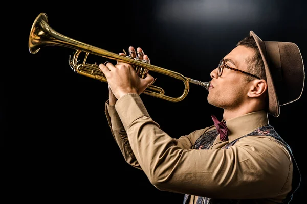 Side view of young male jazzman in hat and eyeglasses playing on trumpet isolated on black — Stock Photo