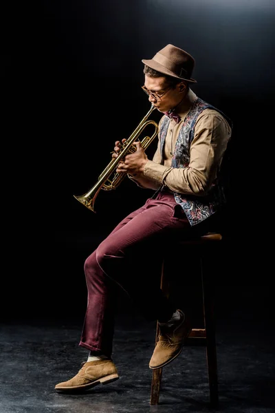 Elegante músico masculino de raza mixta tocando la trompeta mientras está sentado en la silla en negro - foto de stock