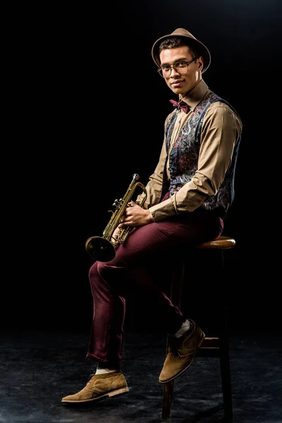 Smiling mixed race male musician posing with trumpet while sitting on chair on black — Stock Photo