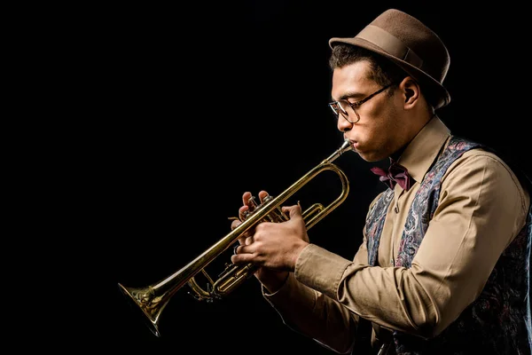 Side view of mixed race male jazzman in hat and eyeglasses playing on trumpet isolated on black — Stock Photo