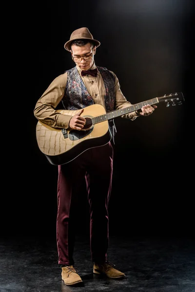 Concentrado joven en elegante sombrero tocando en la guitarra acústica en negro - foto de stock