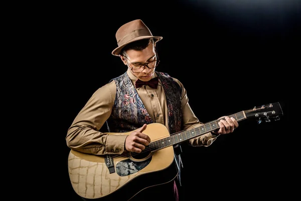 Focused mixed race male musician in eyeglasses and hat playing on acoustic guitar isolated on black — Stock Photo