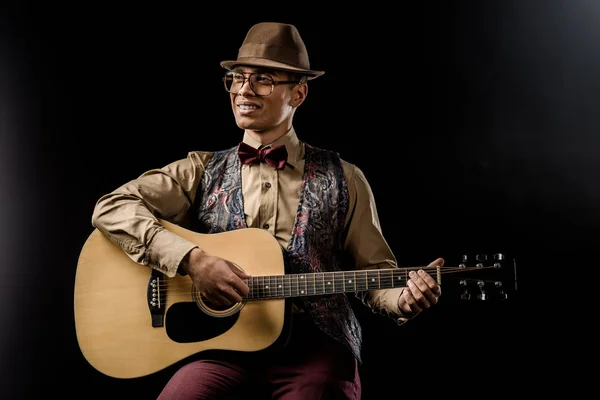 Счастливый музыкант смешанной расы в очках и шляпе, играющий на акустической гитаре, изолированной на черном — стоковое фото
