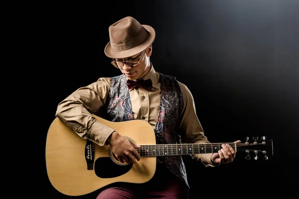 Young handsome man in eyeglasses and hat playing on acoustic guitar isolated on black — Stock Photo