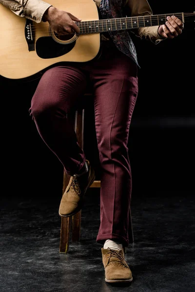 Обрізане зображення молодого чоловіка, який грає на акустичній гітарі, сидячи на стільці на чорному — стокове фото