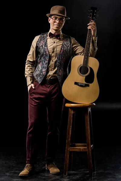 Чоловічий музикант у стильному капелюсі позує з акустичною гітарою біля стільця на чорному — стокове фото