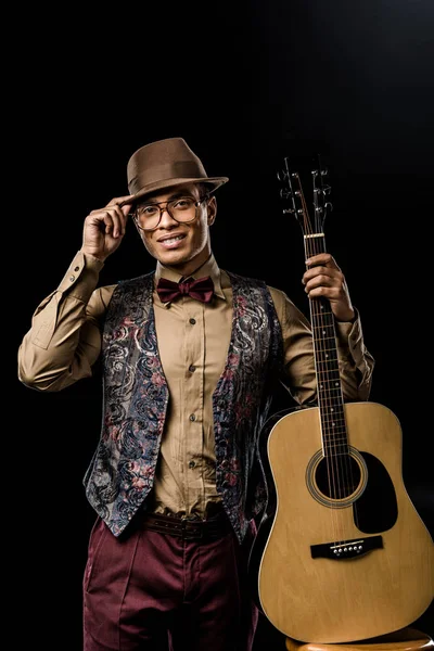 Cheerful mixed race male musician in eyeglasses and hat posing with acoustic guitar isolated on black — Stock Photo