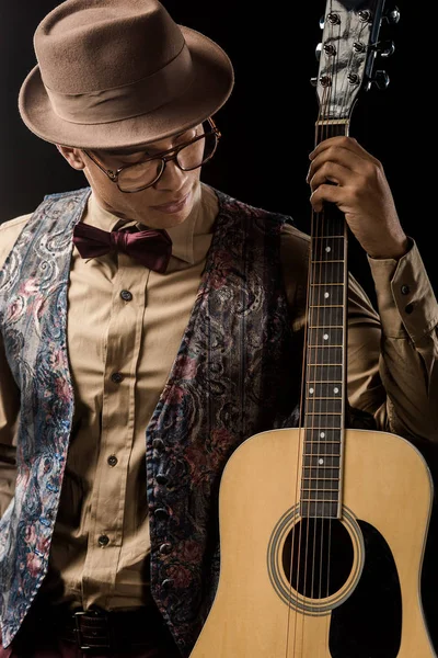 Молодой музыкант смешанной расы в очках и шляпе позирует с акустической гитарой, изолированной на черном — стоковое фото