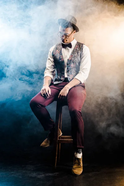 Fashionable mixed race young man posing on chair on stage with smoke and dramatic lighting — Stock Photo