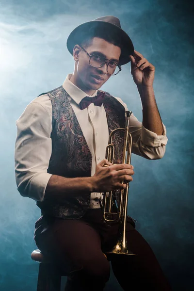 Guapo joven con estilo en sombrero y anteojos posando con trompeta en el escenario con humo e iluminación dramática - foto de stock
