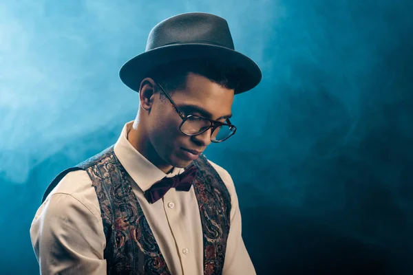 Mixed race young man in stylish hat and eyeglasses posing on stage with smoke and dramatic lighting — Stock Photo
