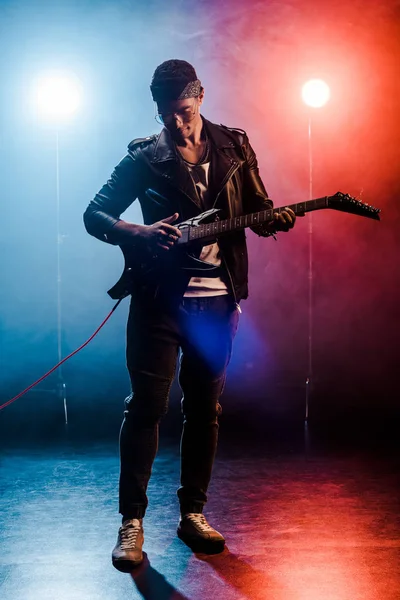 Concentrated male rock star in leather jacket performing on electric guitar on stage with smoke and dramatic lighting — Stock Photo