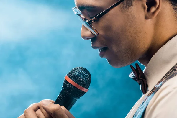 Partial view of mixed race man singing in microphone on stage with smoke and dramatic lighting — Stock Photo