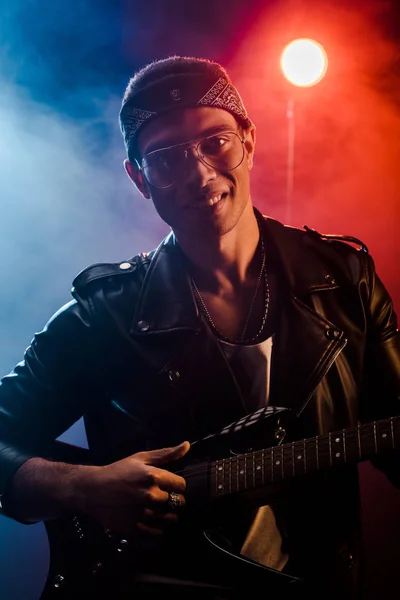 Músico masculino alegre em jaqueta de couro tocando na guitarra elétrica no palco com fumaça e iluminação dramática — Fotografia de Stock