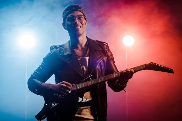 Laughing young male musician in leather jacket performing on electric guitar on stage with smoke and dramatic lighting — Stock Photo