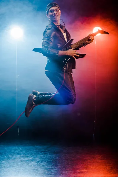 Músico feliz do sexo masculino pulando e tocando na guitarra elétrica durante concerto de rock no palco com fumaça e holofotes — Fotografia de Stock