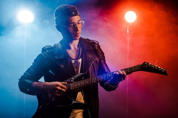 Focused male musician in leather jacket performing on electric guitar on stage with smoke and dramatic lighting — Stock Photo
