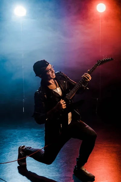 Rock star masculine souriante en veste de cuir jouant de la guitare électrique sur scène avec de la fumée et un éclairage dramatique — Photo de stock