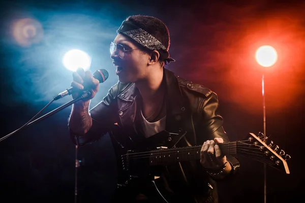 Músico masculino emocional cantando en el micrófono y tocando en la guitarra eléctrica en el escenario durante el concierto de rock - foto de stock
