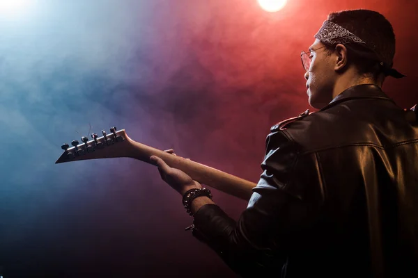 Вид ззаду чоловічий музикант у шкіряній куртці, що грає на електрогітарі на сцені з димом та драматичним освітленням — стокове фото