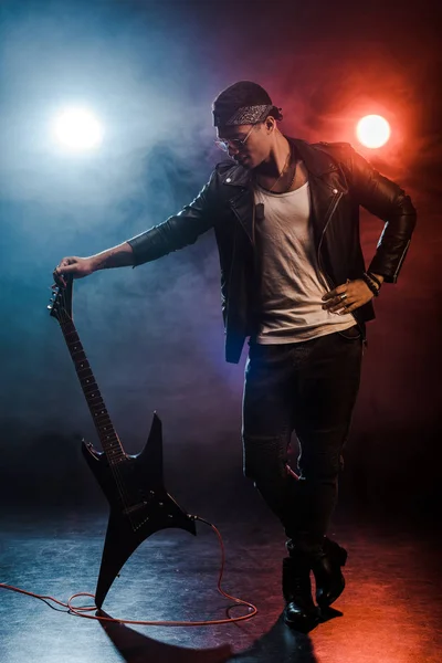 Young mixed race rock musician in leather jacket posing with electric guitar on stage with smoke and dramatic lighting — Stock Photo