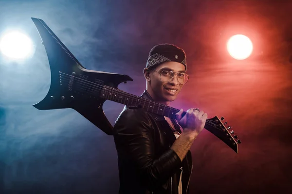 Smiling mixed race rock musician in leather jacket posing with electric guitar on stage with smoke and dramatic lighting — Stock Photo