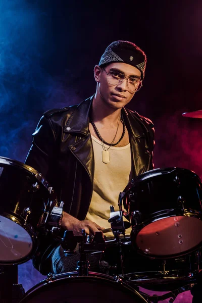 Handsome mixed race male musician sitting behind drum set on stage with smoke and dramatic lighting — Stock Photo